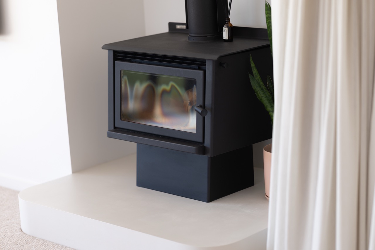 Freestanding wood burner on black plinth above curved microcement hearth with snake plant alongside