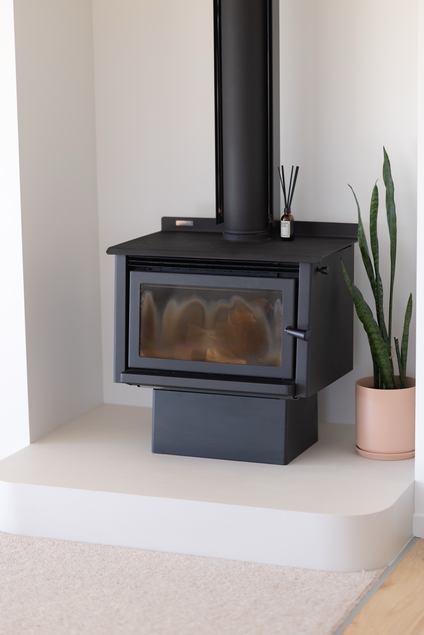 Freestanding black wood burner on plinth above curved microcement hearth, with snake plant in pink ceramic pot alongside