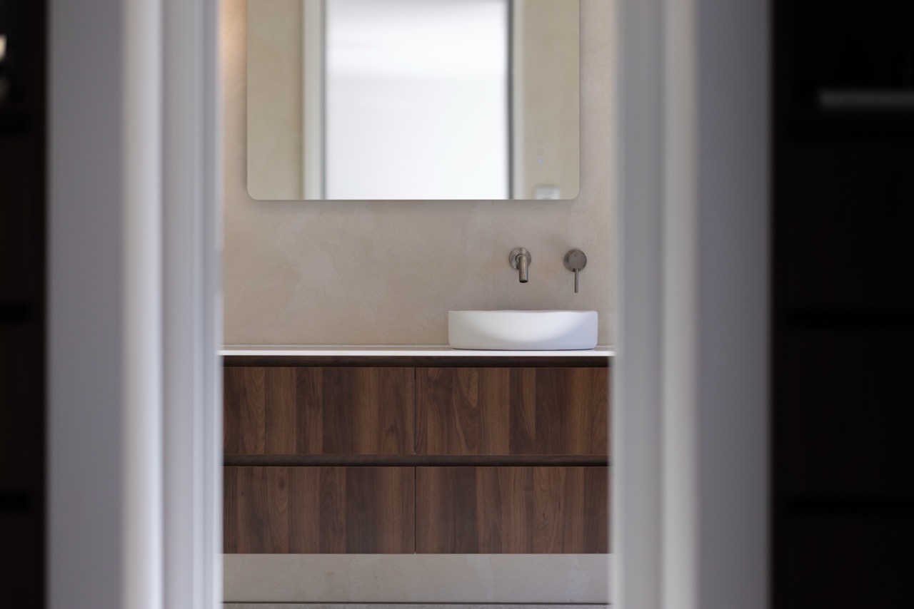 Glimpse through the doorway to the ensuite vanity, walnut drawers, and back-lit mirror