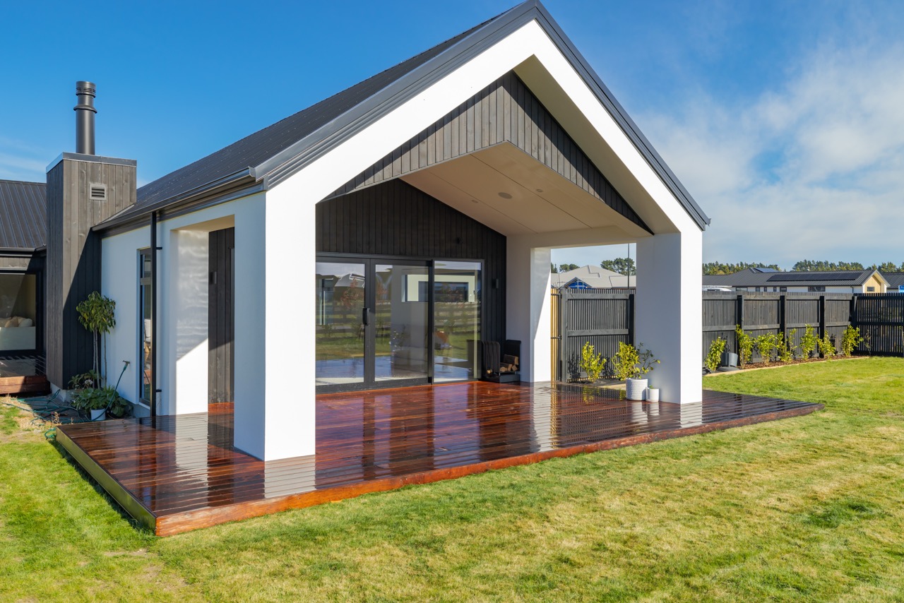 Side angle of rear gable porch with hardwood deck, dark vertical timber, white render columns, and chimney flue