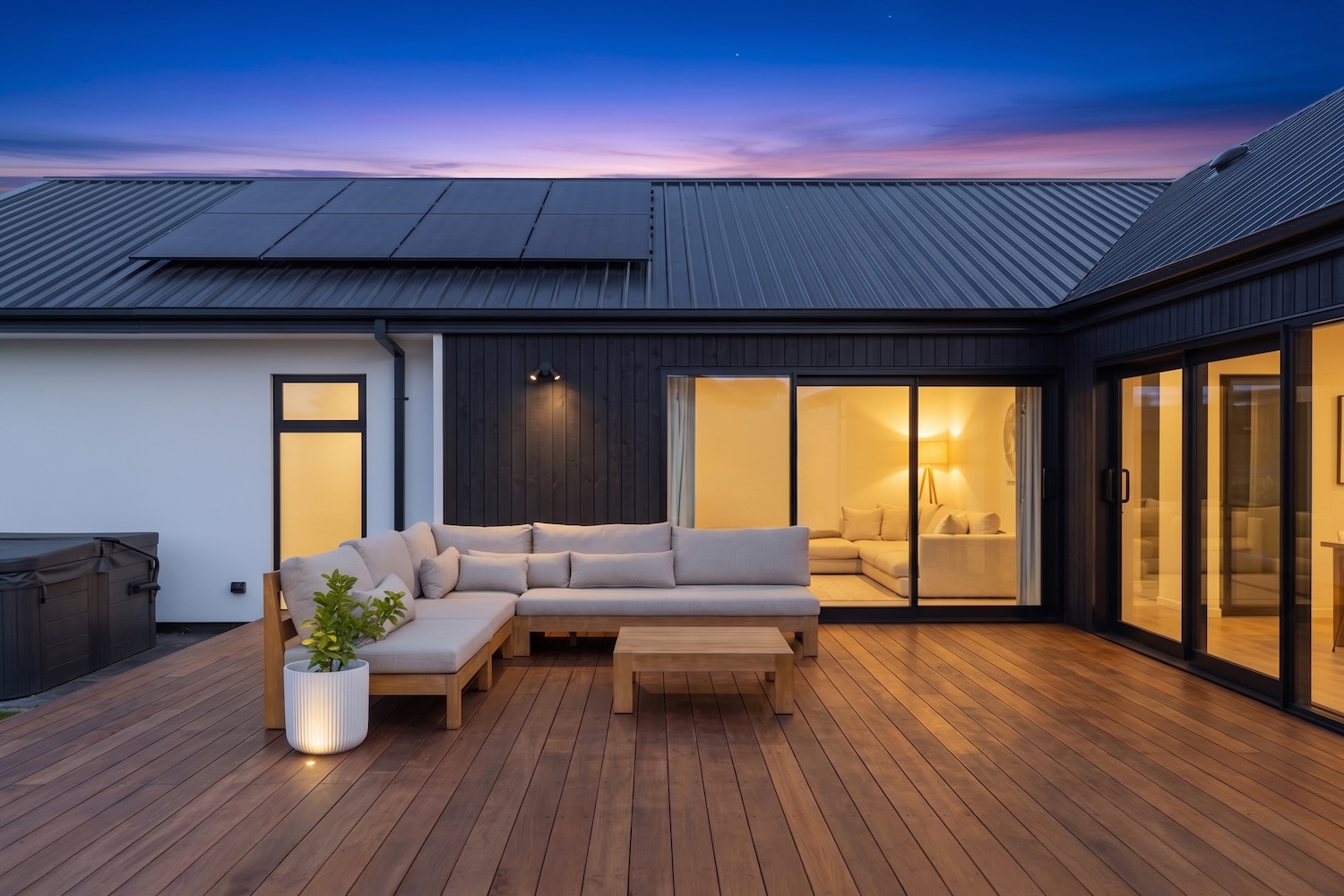 Twilight courtyard at 25 Panama Crescent — outdoor sectional on hardwood deck, dark vertical timber walls, solar-panelled roof, and warm interior glow through sliding doors